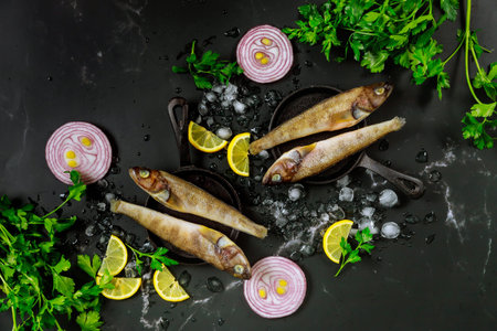 Raw whole fish with ice, parsley and lemon on black table. Top view.