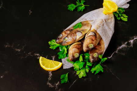 Uncooked whole fish with parsley and lemon wrapped in parchment paper on black table. Top view.
