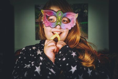 Jewish girl in carnival mask bites hamantaschen cookie on Purim.の写真素材
