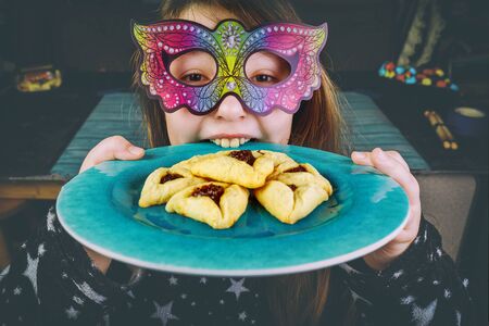 Jewish girl in carnival mask bites plate with purim hamantaschen cookies.の写真素材