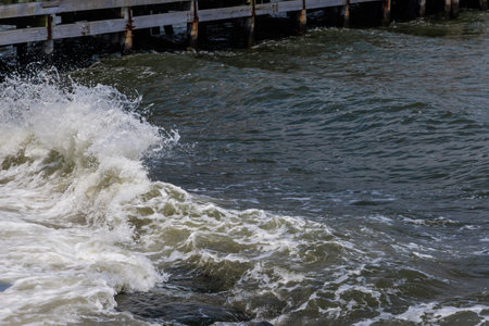 The waves crash vigorously against the wooden pier, sending splashes of water into the air. A cloudy sky looms overhead, adding a dramatic feel to the coastal scene.の写真素材