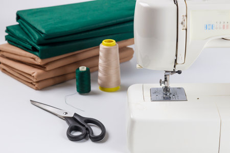 Sewing machine sits on clean surface along with spools of thread and sharp scissors. Neatly folded fabric in green and brown colors awaits crafting and creativity.の写真素材