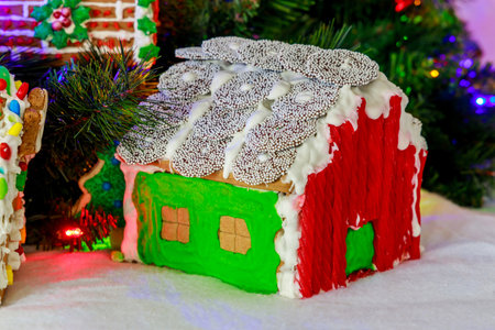 A gingerbread house features vibrant icing with red and green colors and a snowy roof, displayed among Christmas decorations and lights on a cozy table.の写真素材