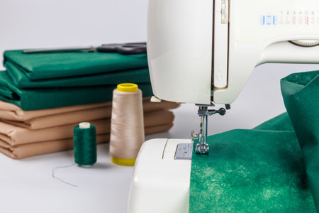 Sewing machine actively stitching green fabric with various spools of thread nearby. Neatly folded brown paper lies on table, indicating preparations for textile projects.の写真素材