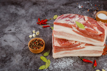 Two thick pieces of pork belly placed on dark surface, surrounded by red chilies, pepper, and aromatic herbs, creating rustic setting for cooking.の写真素材