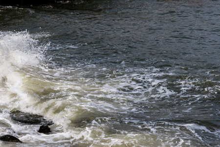 Powerful waves crash against dark rocks along coastal area, creating white foam on water's surface during overcast afternoon. Atmosphere dynamic and tranquil.の写真素材