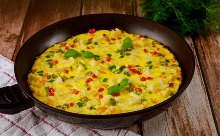 Vibrant vegetable omelette being prepared in black frying pan, featuring colorful bell peppers, herbs, and onions on rustic wooden table with cloth.の写真素材