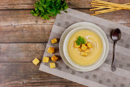 Bowl of creamy soup sits on textured tabletop, garnished with croutons and sprig of parsley. Fresh herbs and breadsticks add touch of elegance to casual setting.の写真素材