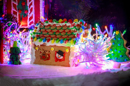 Gingerbread house with bright, colorful roof made of candy and icing sits amidst glowing lights and decorative tree during holiday season.の写真素材