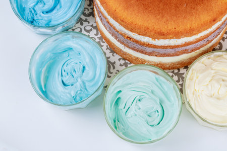 Freshly baked layer cake sits on ornate plate, surrounded by bowls of pastel-colored frosting. Cake ready for decoration with vibrant cream.の写真素材