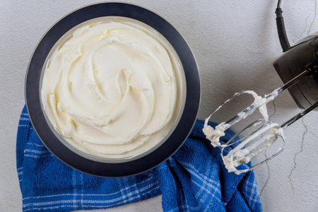Large bowl filled with fluffy whipped cream sits on countertop, accompanied by mixer on blue towel. It looks fresh and ready for use in desserts or toppings.の写真素材