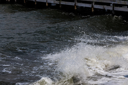 Turbulent waves hit wooden pier, sending splashes of water into air. Cloudy sky casts muted light, enhancing raw beauty of coastal environment.の写真素材