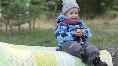 happy little girl drinks tea in the park. The baby is warmly dressed, it is autumn outside. A girl is holding a mug with warm drinks and smiling. Cute videoの写真素材