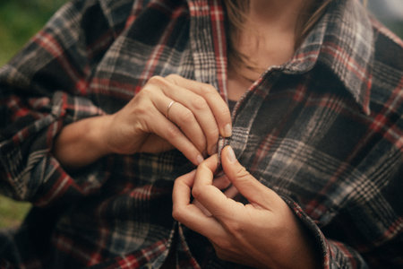 close-up hands buttoning plaid flannel shirt, autumn outdoors, warm sunlight, textured fabric, simple necklaceの写真素材