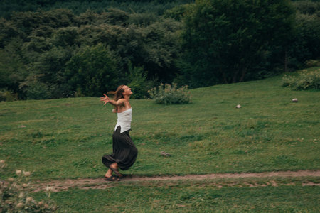 woman walking on grassy hill, carefree stroll through green meadow wearing white top and dark skirt, armsの写真素材