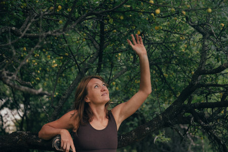 white woman reaching upward toward branch, picking apple in rustic orchard, focused expression, sustainableの写真素材