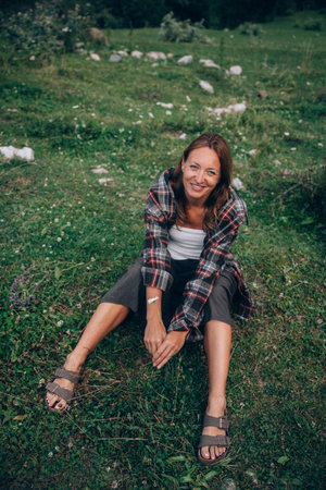 smiling caucasian woman seated on meadow, relaxed travel blogger enjoying green grass, plaid shirt, sandals,の写真素材