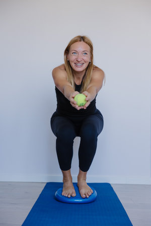 caucasian trainer squatting on balance pad offering tennis ball forward focused smile engaging client-styleの写真素材