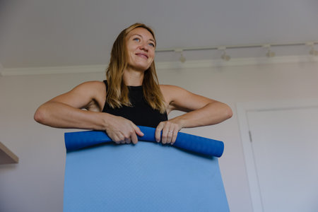 smiling woman holding blue mat preparing yoga session in home studio, fitness coach role, soft natural light,の写真素材