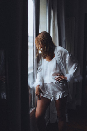 caucasian woman in satin robe standing by window, hands on hips, feather cuff detail, soft morning light,の写真素材