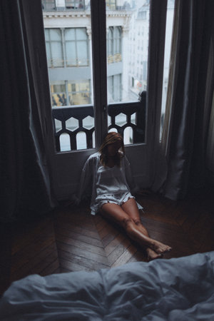 caucasian woman seated on floor looking out over city from tall window, legs extended, back against curtain,の写真素材