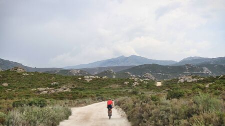 Cycling. Offroad Cycling in Agriates Desert, Corsica, France. a red shirt cyclist, Dusty road, Rocky road, Mediterranean Vegetation. Mountains, Daylight. Cloudy sky. Courage for the Journey.の写真素材