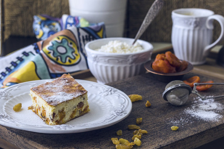 Cottage cheese casserole with raisins and dried apricots. A piece of casserole with powdered sugar on a white plate, fresh farmer cottage cheese in a white bowl on a wooden table. Rustic style.の写真素材