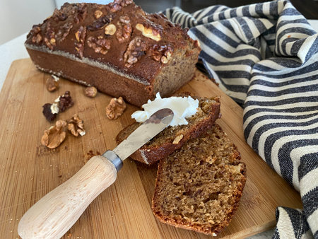Homemade banana bread. Slice cut from a loaf on a cutting board and spread with curd cheese using a...