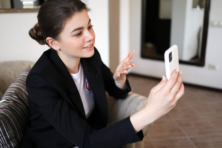 Attractive smiling young woman in a black jacket talking on a mobile video link while sitting in a...
