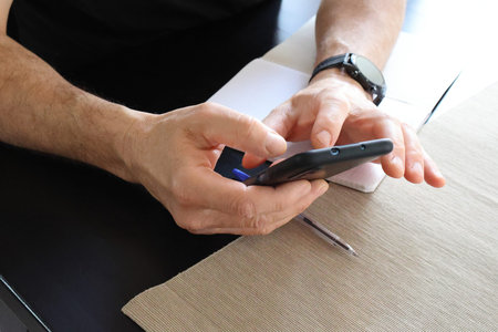 Close-up of the hands of a man with a smart watch on his hand, who is holding a black phone, looking...
