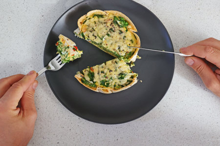 man eats homemade pie with omelette, spinach, red bell pepper with tortilla instead of dough on a dark plate on the table, healthy breakfast or brunch. View from above.の写真素材