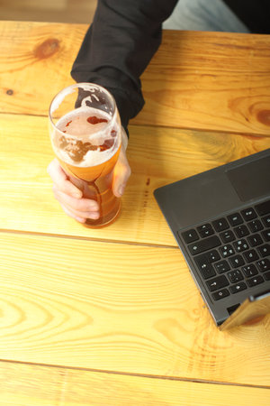 mug with beer in hand next to laptop on wooden tableの写真素材