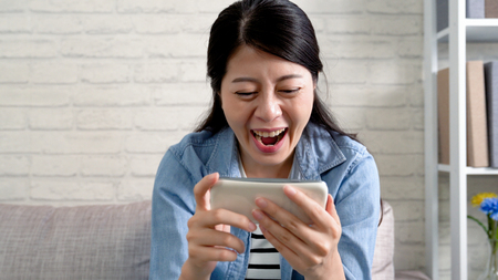cheerful woman laughing watching football game on cellphone. female fan love soccer match looking online on smartphone at home in comfortable sofa. young lady smiling happily spending time indoors.の写真素材