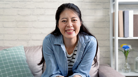 happy asian woman sitting on comfortable couch cheerfully smiling face camera. young housewife enjoy love her life concept. joyful lady at home having fun laughing.の写真素材