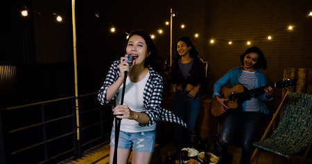 three asian love playing music on the roof. sexy singer holding microphone performing facing camera smiling singing. young ladies in the background dancing laughing.の写真素材