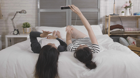 two asian teenage girls lying on bed upside down holding cellphone having video phone call via smartphone.の写真素材