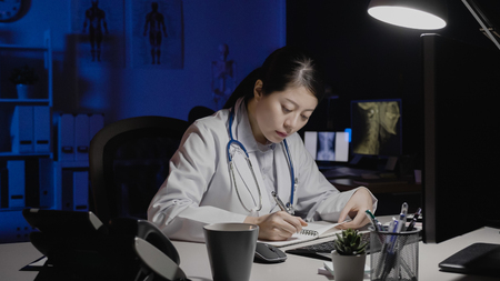 asian female doctor writing on notepad in medical office.の写真素材