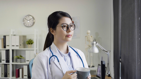 Young chinese asian female doctor having a break and drinking coffee watching outdoor urban view from sunshine window.の写真素材