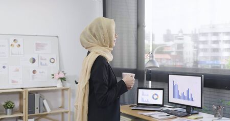 side view adult muslim businesswoman ceo standing at desk in modern office with cup of coffee. elegant arabic woman looks out of window and contemplates the city and skyscrapers. tea break lifestyle.の写真素材