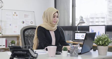 Young islam female employee looking at computer monitor during working day in office typing on laptop keyboard. elegant positive muslim lady manager in suit sitting workplace desk with morning coffeeの写真素材