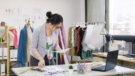 Young people small business. asian woman at work as fashion designer and tailor looking at sketches of new collection in atelier on desk. dressmaker lady in eyeglasses concentrated in studio workshopの写真素材