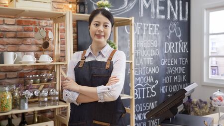 successful asian businesswoman happy cafeteria owner looking at camera smiling. confident young woman barista wearing apron welcoming guests with crossed arms. prosperous catering business concept.の写真素材