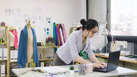 Responding on business e mail. Beautiful young Asian chinese woman working using computer and smiling while standing in workshop. fashion designer discussing with customer customize dress making.の写真素材