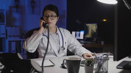 asian female doctor wear eyeglasses answer phone call while using computer working in dark clinic office at night.の写真素材