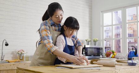 asian korean mother and daughter in apron using hands not rolling pin kneading dough for cookies together in kitchen on easter holidays. young mom teaching little girl baking cake on mothers day.の写真素材
