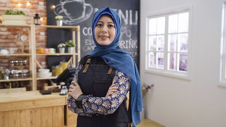 portrait of attractive asian muslim female cafe startup business owner standing crossed arm in coffee shop.の写真素材