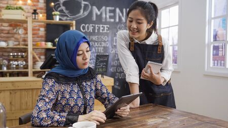 Portrait of smiling young japanese waitress taking order from malay client in cafe bar writing note. muslim woman guest using digital tablet reading menu while friendly female barista introduceの写真素材