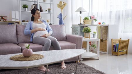 asian japanese pregnant woman sit on grey sofa with her hand on her stomach. Waiting for the birth of unborn baby in belly. young elegant lady in maternity relax in couch in living room at home.の写真素材
