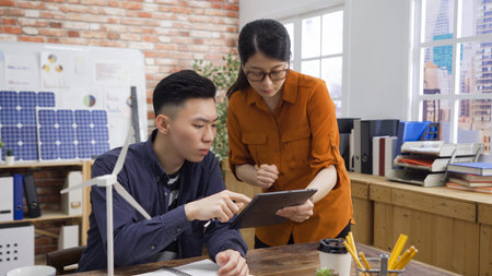 confident asian woman engineer asking opinion of her male colleague in modern office with red brick...
