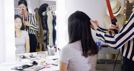 asian japanese girl professional hairdresser curling model hair stand behind on backstage. concept of hairstyle preparation. mirror reflection of young lady celebrity relax sitting at makeup table.の写真素材
